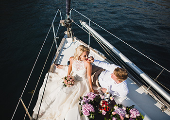 picture from the top of the boat of a newly married couple on the top of a boat while sailing through the water and there are purple flower bouquets in the scene